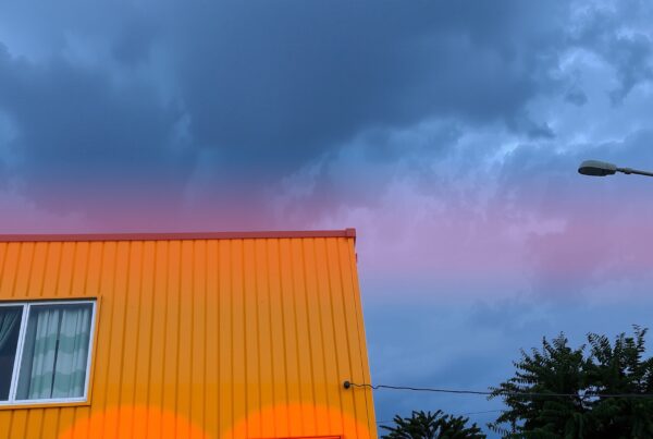 Vivid yellow building with corrugated metal exterior set against a dramatic sky, showcasing pink and purple cloud formations, with streetlamp and trees.