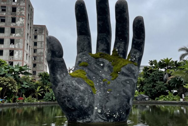 A giant hand sculpture emerges from a reflective pond amidst lush greenery and an old building with overcast skies.