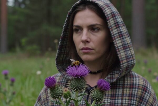 A contemplative woman in a hooded coat stands in a green field with a bee on a purple thistle flower.