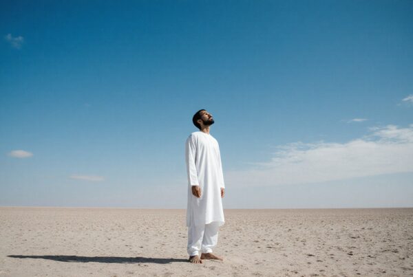 A man in white clothing stands barefoot in a vast desert, gazing thoughtfully at the open blue sky.
