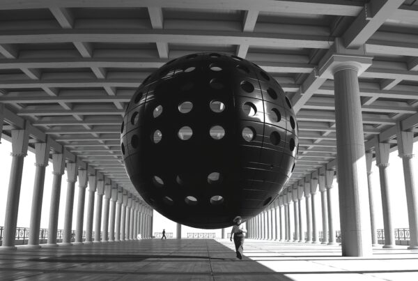 A large black sphere with circular holes is suspended in a classical colonnade, blending modern art with traditional architecture.