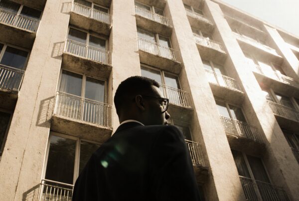 A man in a suit gazes thoughtfully up at a tall building in an urban setting.