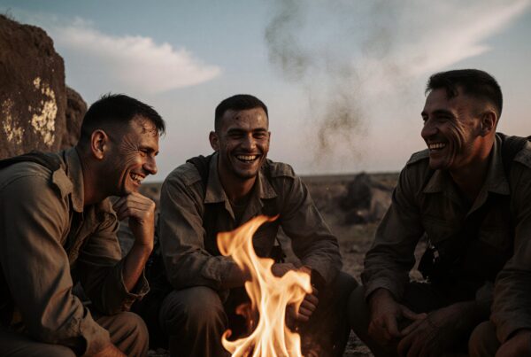 Four soldiers laughing around a campfire in a rugged outdoor setting.