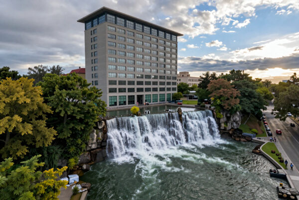 Modern building beside scenic waterfall with walking paths and parked cars during sunset
