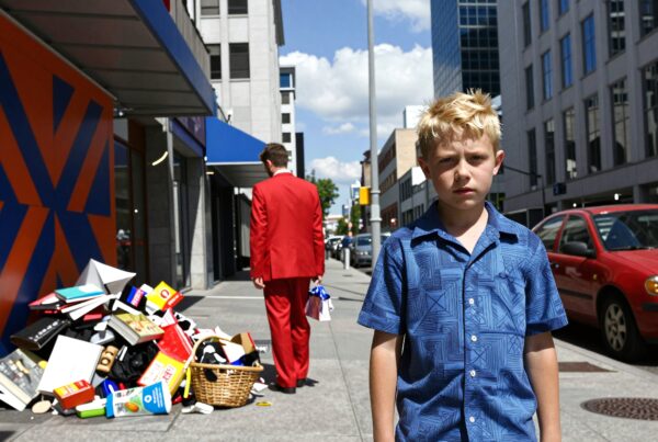 A young boy stands on a city sidewalk with piles of books and a man in a red suit.