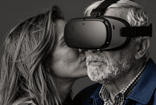 Elderly man wearing VR headset kissed by loving partner embracing futuristic technology