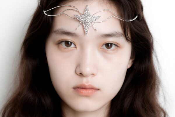 A young woman with dark hair and a sparkling star-shaped ornament on her forehead gazes calmly at the camera against a soft white background.