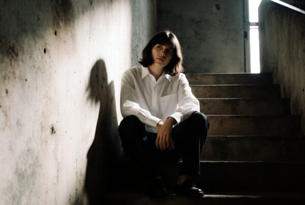 Young person in white shirt sits pensively on dimly lit urban staircase, casting shadows on textured wall.