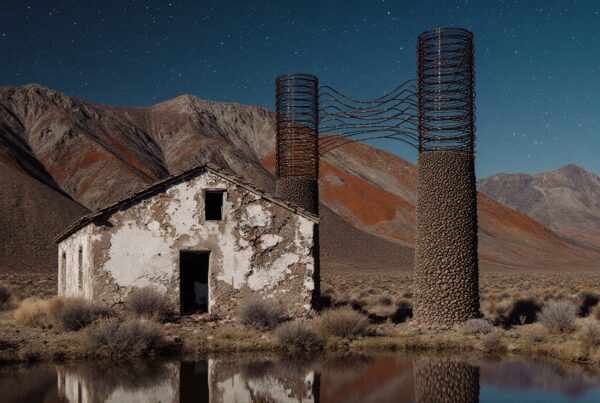 Abandoned building with stone towers in desert at night reflecting in water, with stars and mountains.