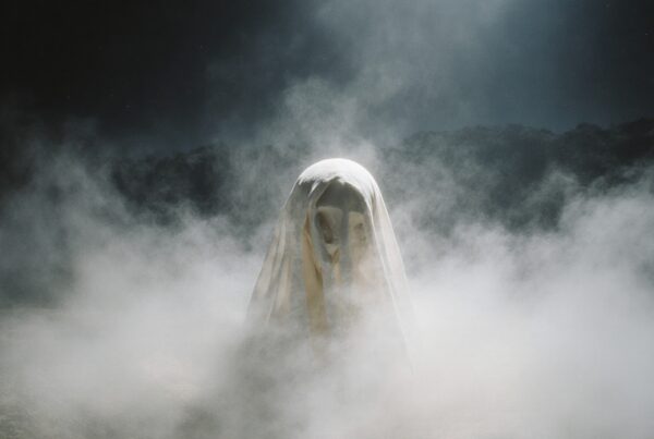 A ghostly figure shrouded in white stands in an eerie mist-covered landscape at night.
