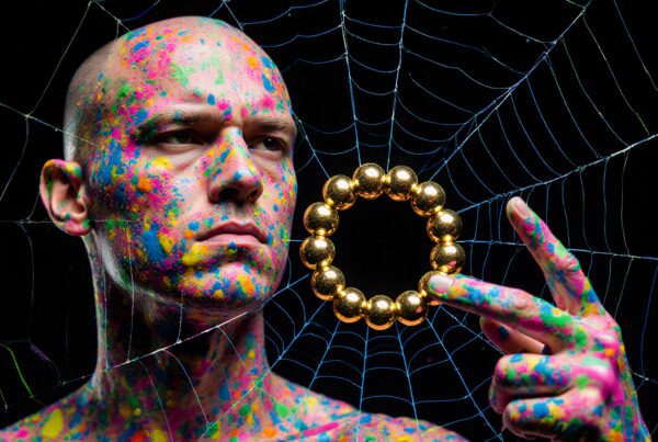 A bald man with colorful paint holds a gold ring amidst a spider web.