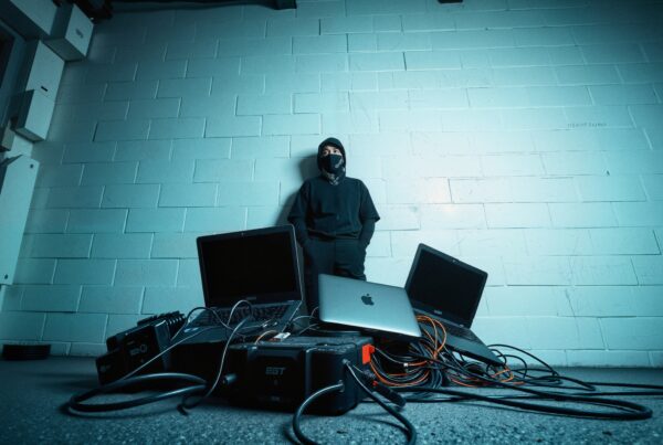 Person in dark clothing amidst laptops and cables, suggesting secretive tech activity.