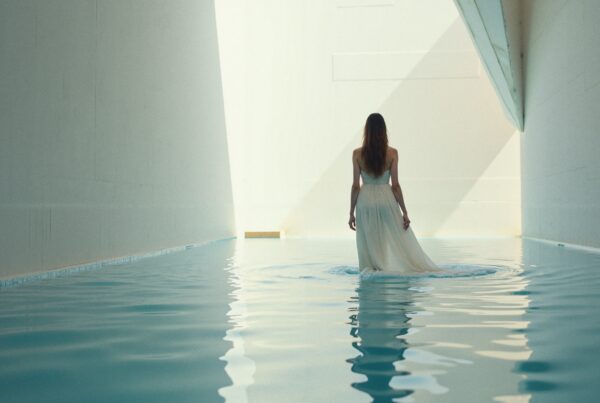 A woman in a flowing white dress stands in shallow water, facing the light in a serene, minimalist setting.