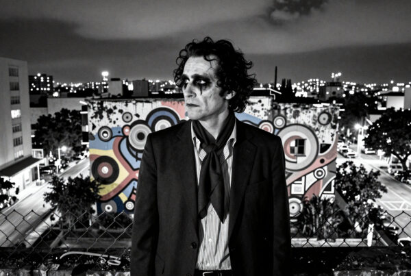 Actor dressed as Joker standing alone atop rooftop against nighttime city backdrop with colorful mural behind