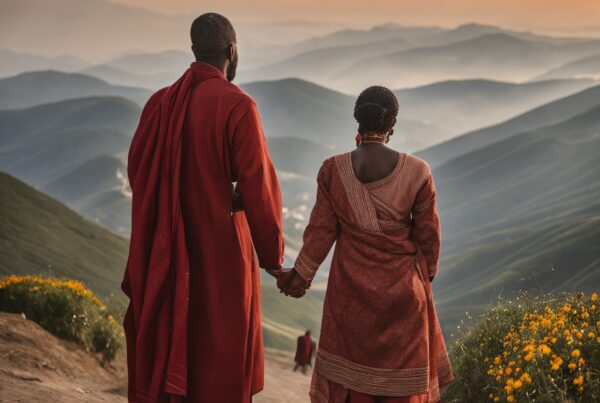 A couple in red traditional attire holds hands on a scenic mountain path at sunset.