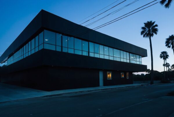 Image of a modern building with large windows reflecting the twilight sky, framed by palm trees and power lines.