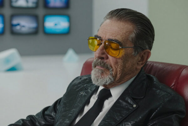 Elderly man wearing sunglasses sitting in a red leather chair surrounded by computer monitors