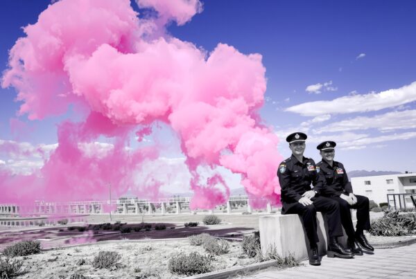 Two officers seated outdoors with a vivid pink smoke cloud behind them under a blue sky at a formal event.