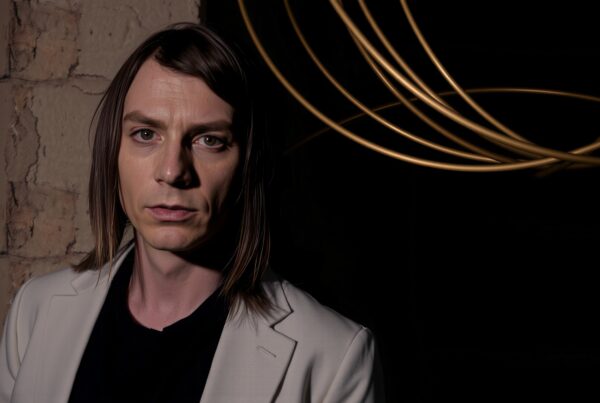 A person with long brown hair in a blazer, standing against a textured wall with gold decorative rods.
