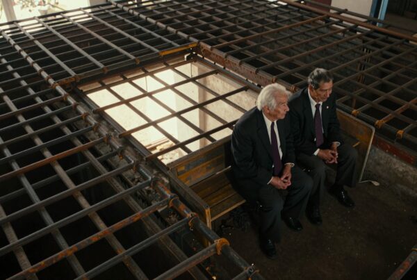 Two men in formal suits sit pensively inside a metal-bar structure, sunlight filtering through.