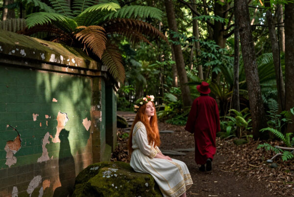 Woman in floral crown sitting peacefully near mossy stone in sunlit forest glen wearing ornate white gown as cloaked figure walks away along winding dirt trail surrounded by tall ferns and decaying painted walls