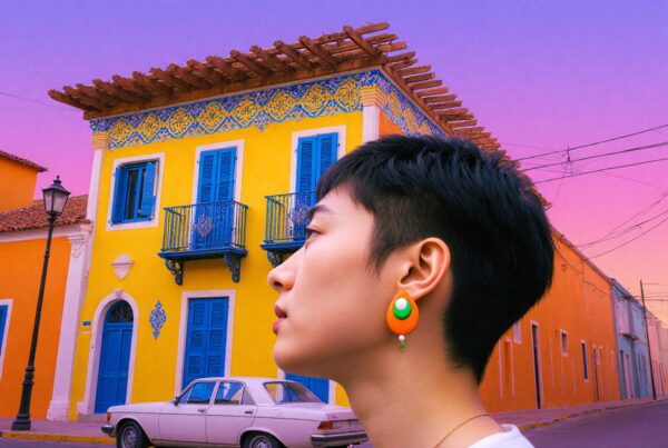 A profile view of a person wearing a colorful earring stands against a vibrant yellow building with blue accents during a pastel sunset.