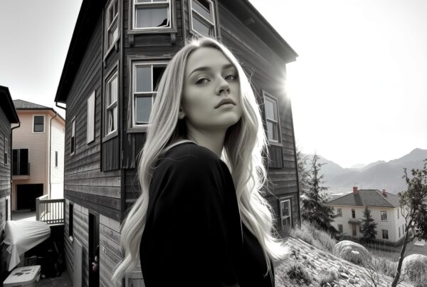 A woman stands near a wooden suburban house, gazing thoughtfully with mountains in the background.