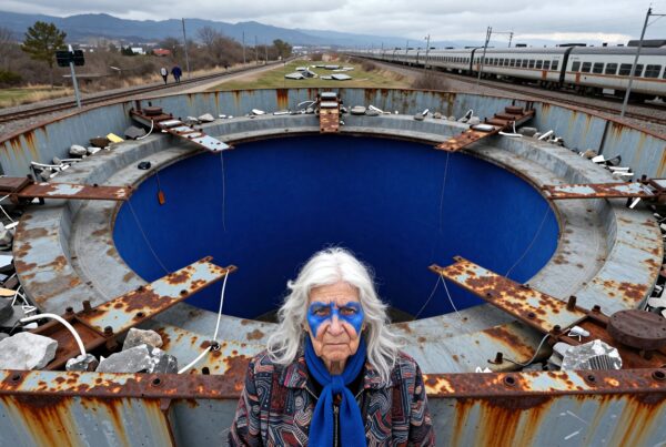 A person with blue face paint stands by a circular metal structure near train tracks.