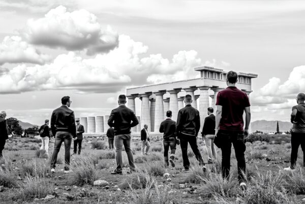Black and white photo of people visiting an ancient Greek temple with dramatic clouds in the background.
