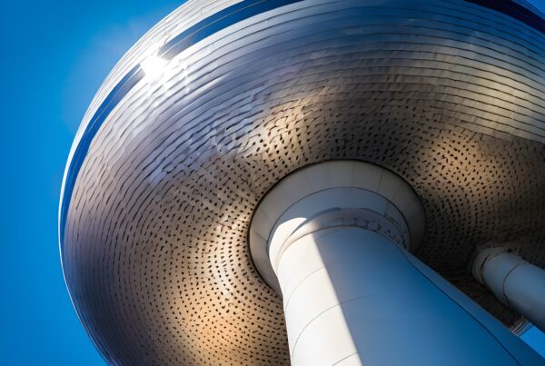A futuristic water tower with metallic finish reflecting sunlight against a clear blue sky, supported by cylindrical pillars.