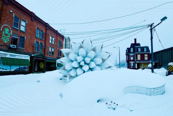 A large geometric snow flower sculpture in a snowy urban street with historic buildings.