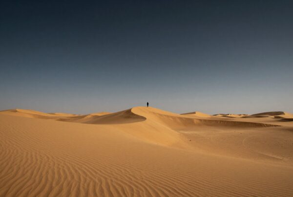 Lone figure on vast desert sand dune under clear sky.