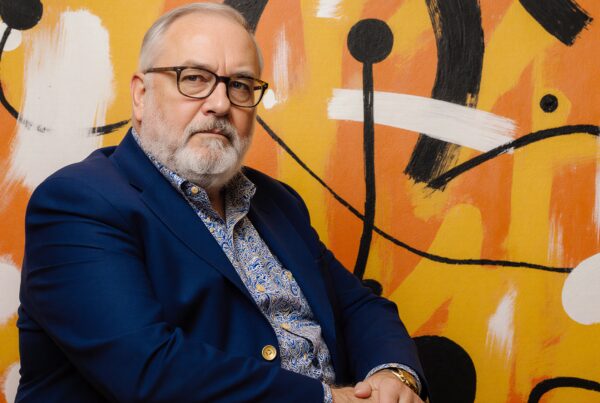 A contemplative elderly man sits thoughtfully against a vibrant abstract mural background, wearing a blue blazer and patterned shirt.