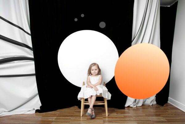 A young girl in a white dress sits calmly on a wooden chair against a backdrop of black and white circles with an overlapping orange element, on a polished wooden floor.