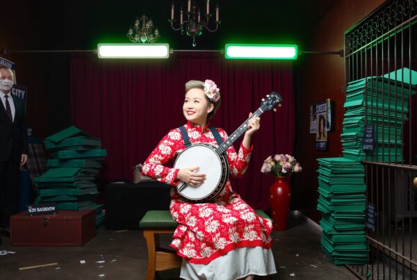 A woman in a red floral dress plays a banjo in an eclectic, theatrical setting, with a man in a suit nearby.