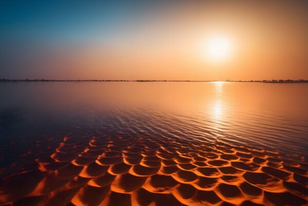 A tranquil sunset casts a golden glow over calm waters with intricate sand patterns in the foreground.