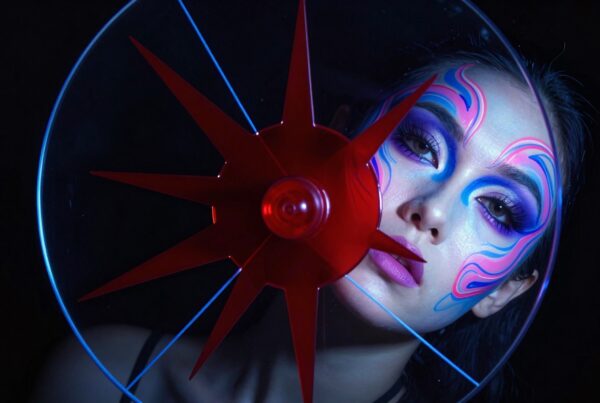 A person with intricate blue and pink makeup looking through red star-shaped art against a dark backdrop.
