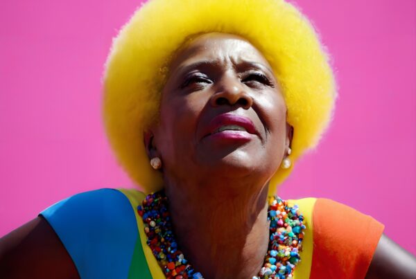 Elderly woman with yellow afro, colorful beads, vibrant background.