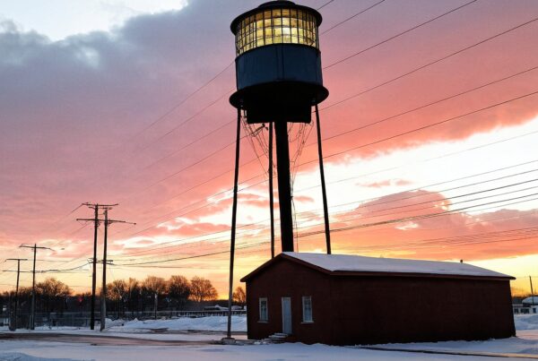 A water tower silhouettes against a vibrant sunset, with snow-covered tracks and a brick building below.